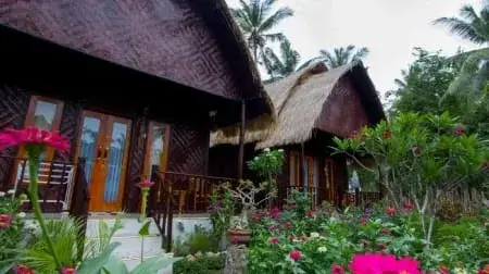 努沙卜尼达（Nusa Penida）度假木屋，茅草屋顶和阳台被热带花园环绕，前景有盛开的粉色花朵。