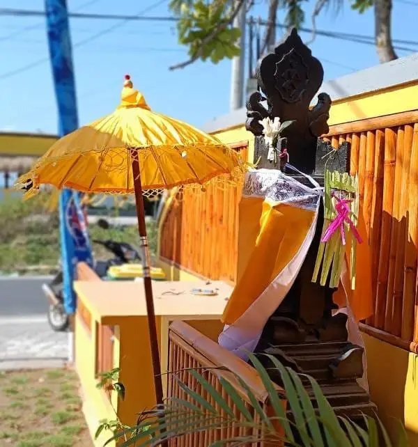 巴厘风格神像被黄白飘带和供品装饰，旁边撑着黄色仪式伞，画面在Nusa Penida路旁寺庙入口举行祭祀场景。