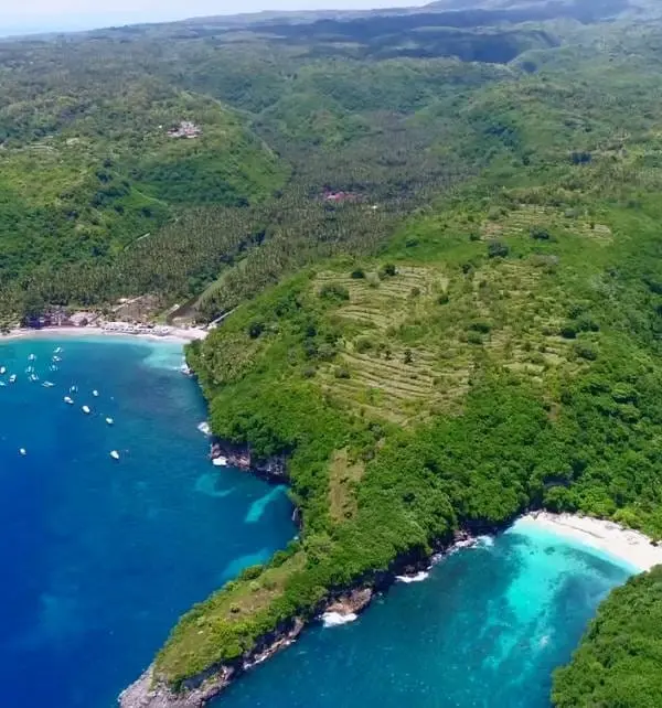航拍Nusa Penida海岸景色，游客船只停在海湾，翠绿山坡與白沙海滩下的清澈湛蓝海水形成对比。