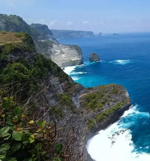 悬崖边观景点俯瞰Nusa Penida海岸,碧蓝海浪拍打白色岩岸,几座独立岩礁在远处海面上显眼地矗立。