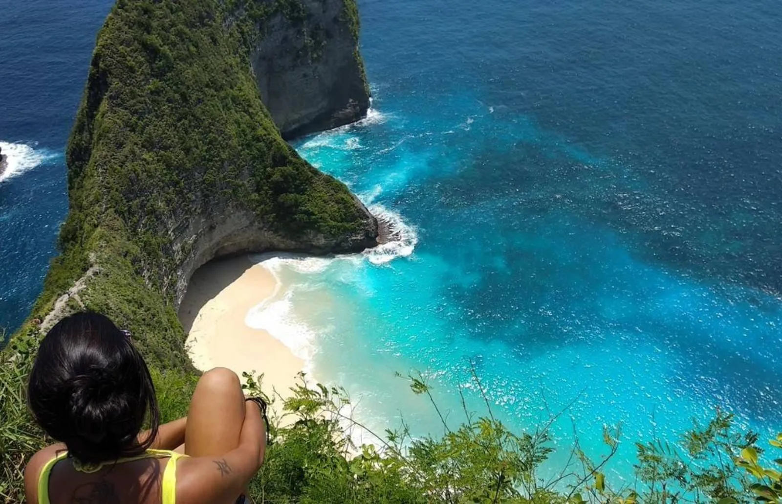 一名游客坐在悬崖边俯瞰Nusa Penida碧蓝海湾和弯曲白沙海滩，阳光下海水呈渐变蓝色。